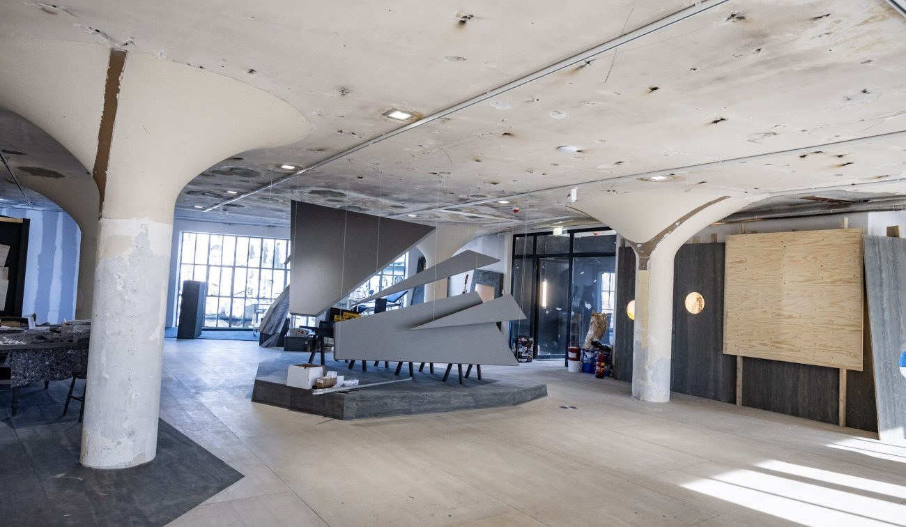 Interieur Nederlands Mijnmuseum in verbouwing met paddenstoelkolommen, open betonnen plafond en veel daglicht; in het midden een groot grijs, hoekig tentoonstellingsobject op een verhoogd plateau, bouwmaterialen tegen de wanden.
