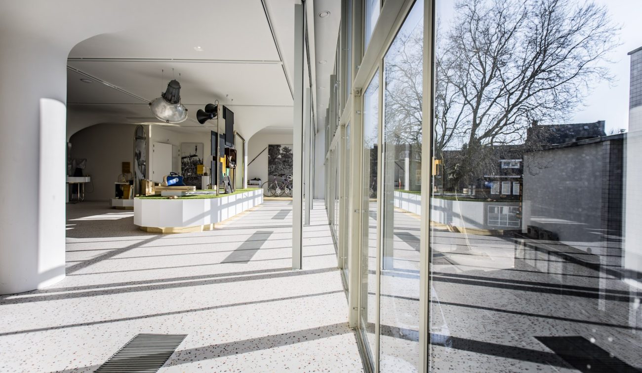 Lichte museumruimte met grote glazen gevel en lange schaduwen op een terrazzo vloer; in het midden een lang wit vitrinedisplay met mijngereedschap. Buiten zijn bomen en gebouwen zichtbaar.