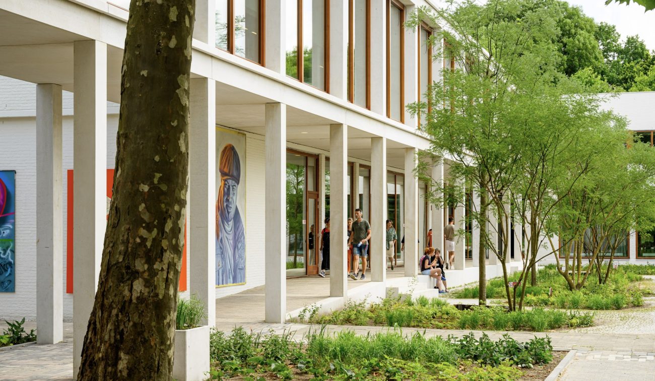 Overdekte looproute langs glazen puien; leerlingen pauzeren tussen jonge bomen en borderplanten.