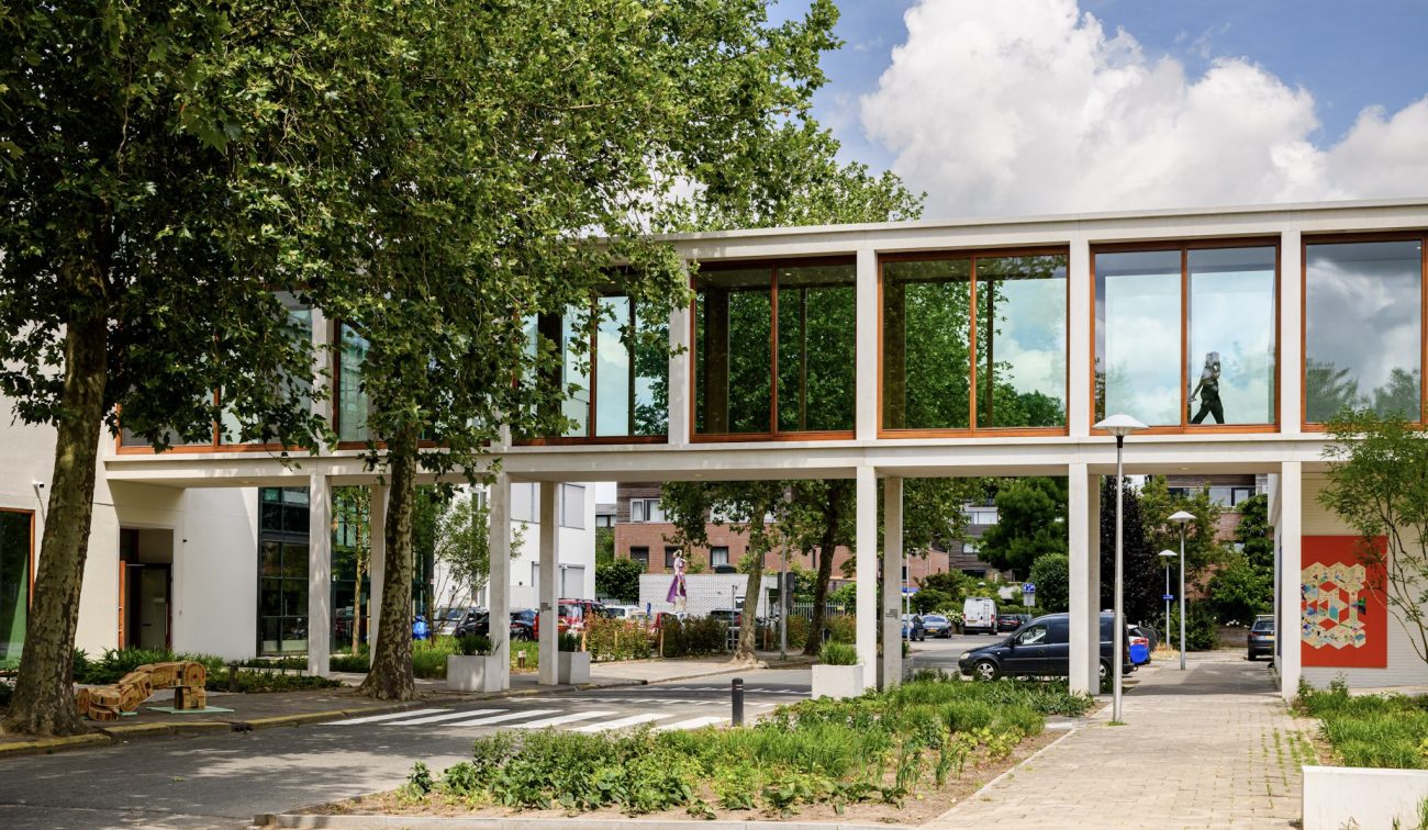 Glazen loopbrug op slanke kolommen met houten kozijnen, tussen bomen en parkeerterrein.