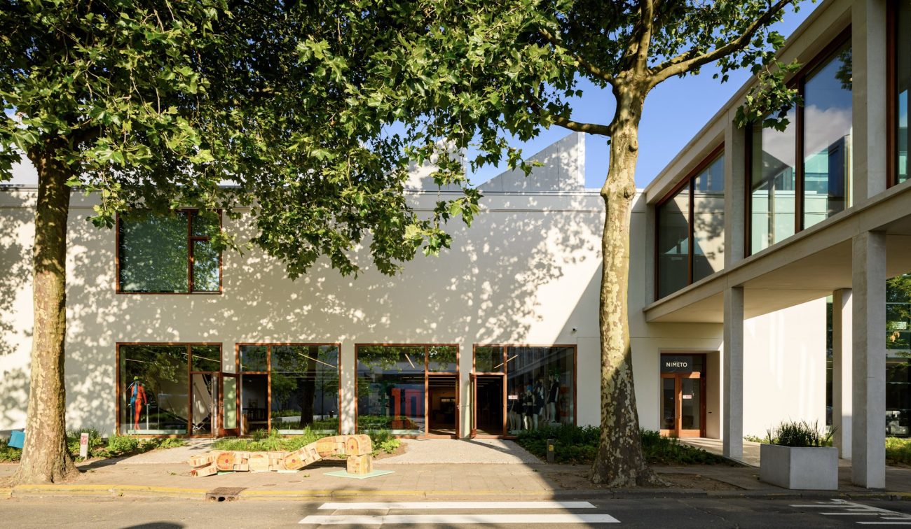 Moderne school met houten accenten, open deuren en overdekte doorgang rechts met “NIMETO”-bord.