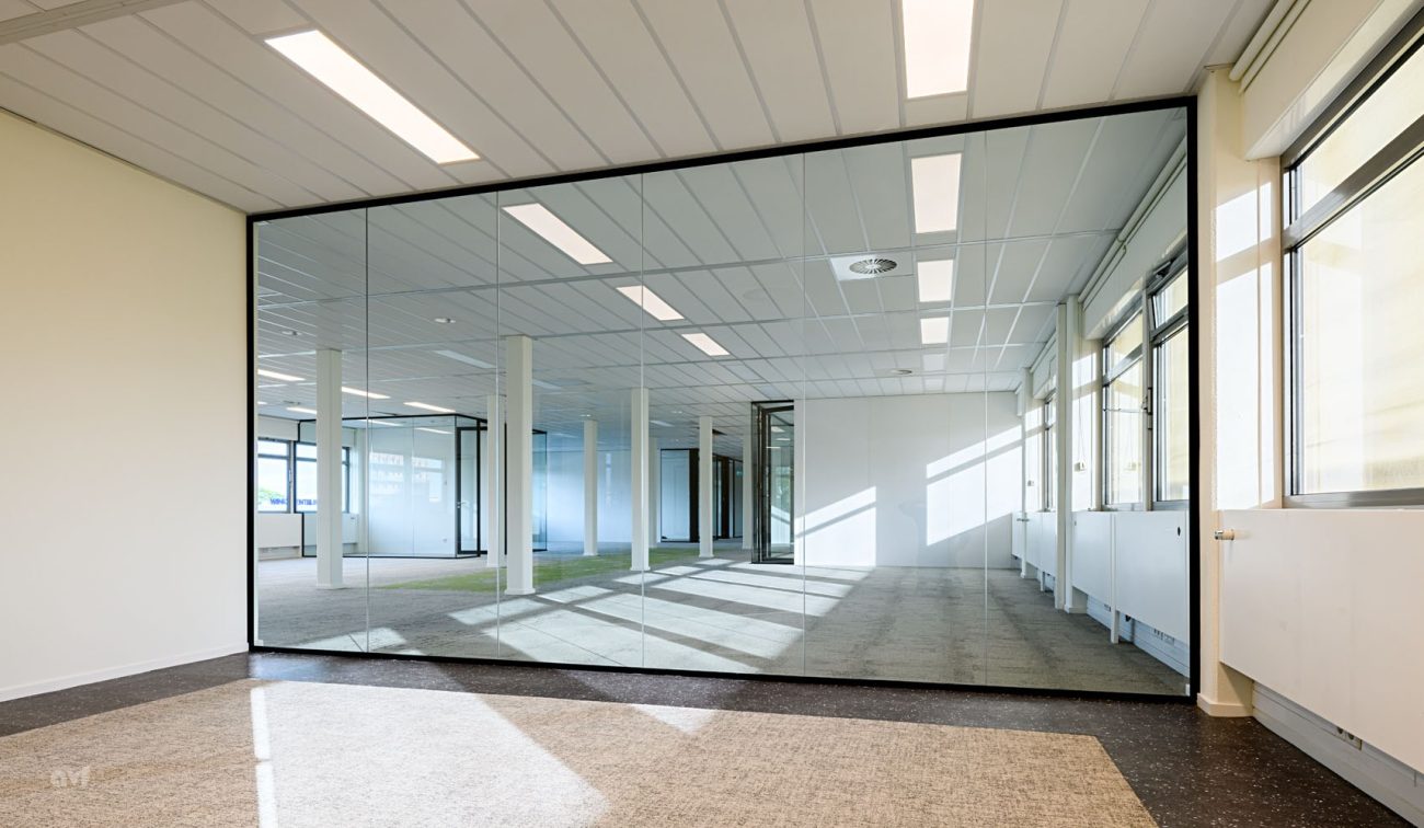 Opgefriste werkvloer met glas/spiegelwand, kolommenrij en grijze tapijttegels in zonlicht