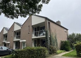 Rij woningen in Stramproy (groot onderhoud) met bakstenen gevels, zadeldaken en witte geveldelen; woningen hebben balkons met glas/white hekwerk, klimplanten langs de gevel en een groen voorterrein onder grijze lucht.