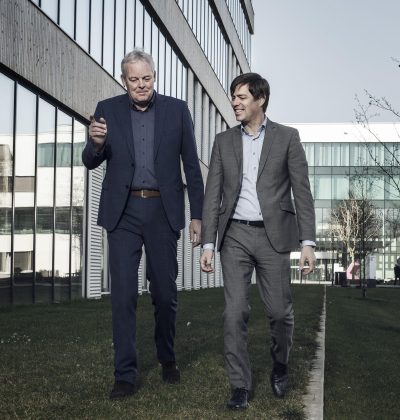 Twee mannen in pak lopen en praten samen langs een modern kantoorgebouw.