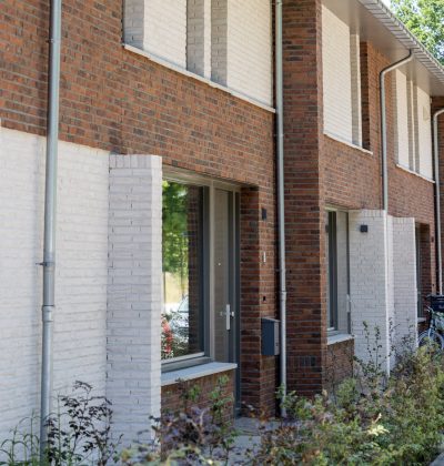 Close-up van een rij nieuwbouwwoningen met rode en wit geschilderde baksteen, grote ramen en regenpijpen; kleine voortuinen met jonge beplanting en een fiets bij een entree.