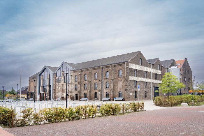 Gerestaureerde Greswarenfabriek in Reuver met bakstenen gevels, grote glazen entree en historische gevelletters; voorliggend plein met fietsrekken, bankjes en jonge bomen.