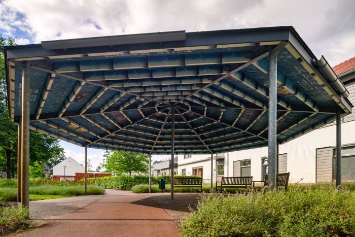Overdekte ontmoetingsplek met kunstzinnig blauw dak, bankjes en groene wandelpaden tussen witte woningen in Kloostervelden, Sterksel.