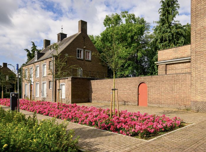 Transformatie Mariakerk Valkenswaard – monumentale pastorie met kloostermuur en rode deuren, langs bloeiende roze perkplanten en jonge leibomen op het binnenplein.