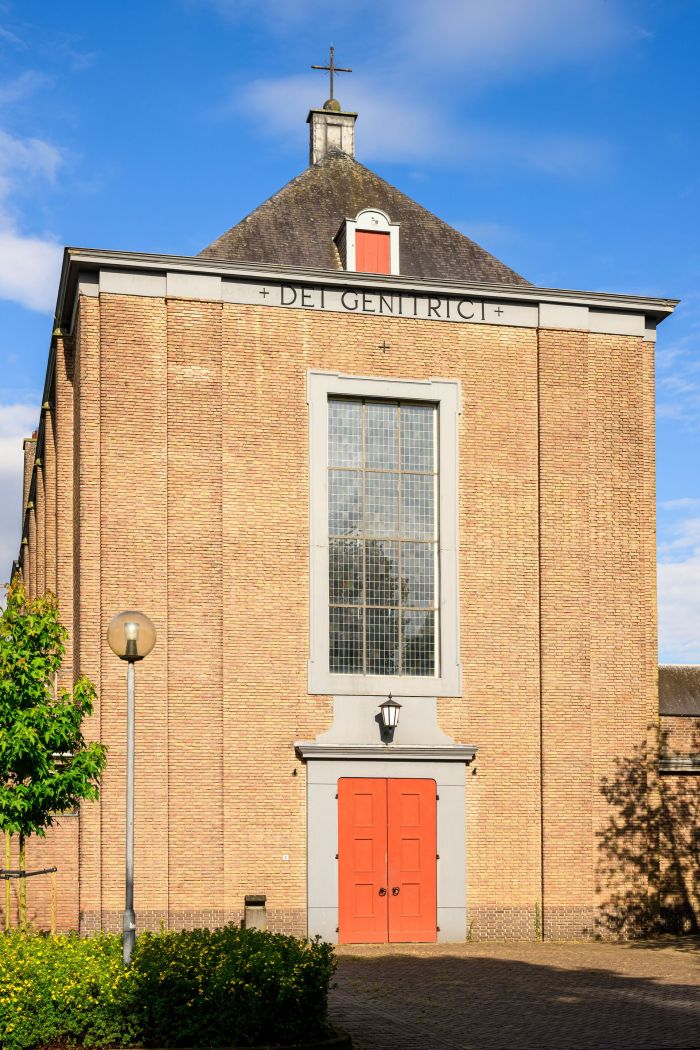 Mariakerk Valkenswaard voorgevel – hoge bakstenen wand met centraal glas-in-loodraam, rode dubbele entree en opschrift “DEI GENITRICI”, onder kap met kruis in de zon.
