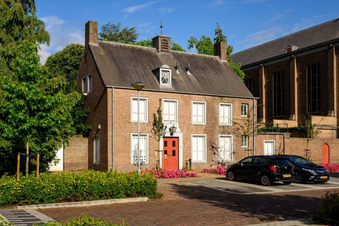 Pastorie bij Mariakerk Valkenswaard met rode voordeur en bakstenen gevel, dakkapel en kerk op de achtergrond, in zomerzon met bloemperk en geparkeerde auto’s.