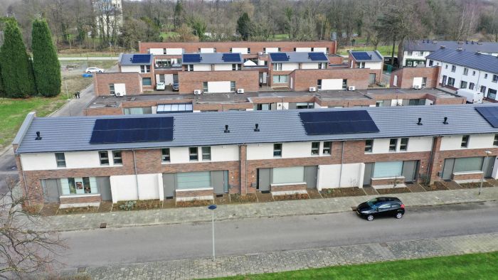 Oblique luchtfoto van nieuwe rijwoningen in Nazareth (Maastricht) met wit-bakstenen gevels en grijze zadeldaken met grote rijen zonnepanelen; daarachter meerdere blokken met platte daken. Een geparkeerde auto staat langs de straat.