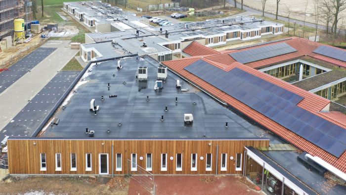Luchtfoto nieuwbouw zorgwoningen Amstenrade: paviljoen met houten gevel, zwart plat dak met installaties, naast rood pannendak met grote zonnepanelen.