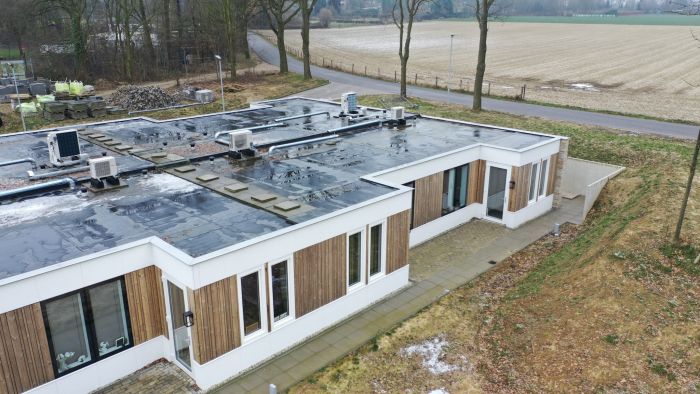 Hoekaanzicht nieuwbouw zorgwoning Amstenrade met witte en houten gevel, plat dak vol installaties, betegeld pad langs de gevel en landelijke omgeving met weg en velden.