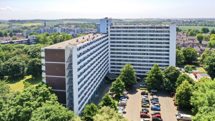 Luchtfoto van L-vormige galerijflat Veliahof in Maastricht met witte gerenoveerde gevels en lange balkonstroken, omringd door bomen en parkeerplaatsen.