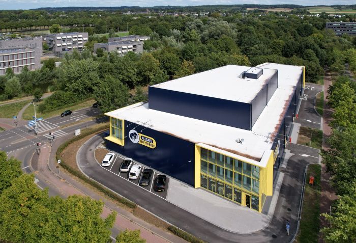 Luchtfoto van de nieuwbouw van Allsafe Self Storage in Maastricht: donkerblauw gebouw met witte dakplaat en felgele kaders rond de glazen hoekgevel, kleine parkeerstrook met auto’s langs de gevel, gelegen aan een kruising en omringd door bomen.