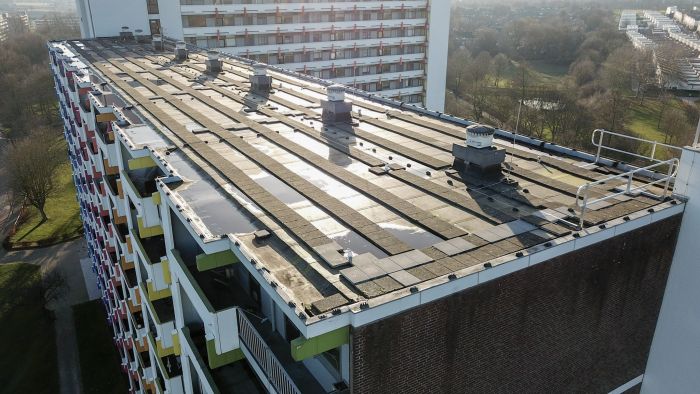 Luchtfoto van het platte dak van een galerijflat met bitumenbanen, ventilatie-units en valbeveiligingsrailing; gekleurde balkonpanelen aan de gevel.