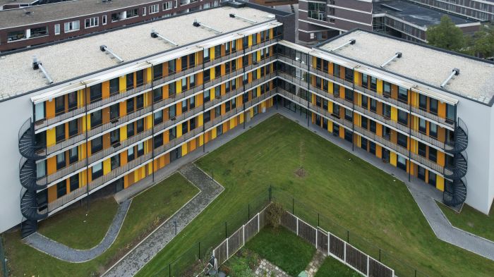 Luchtfoto van nieuwbouw Magistraat Heerlen: U-vormige blokken met doorlopende balkons, gele en oranje panelen en een centrale groene binnentuin.”