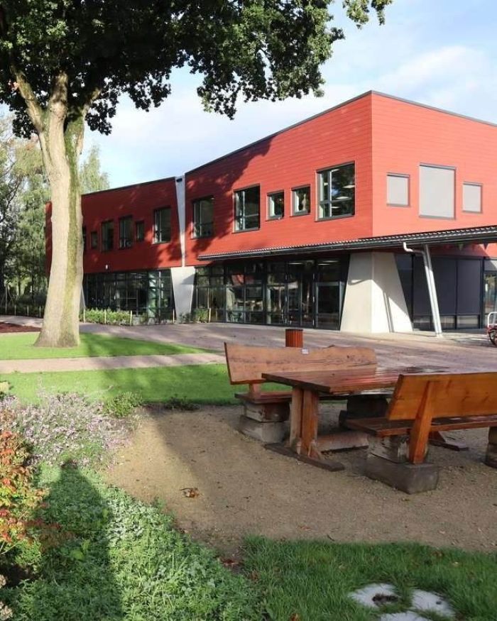 Nieuwbouw van basisschool Den Doelhof in Meijel met rood beklede gevel en grote ramen; voor het gebouw liggen houten picknickbanken op het groene schoolplein.