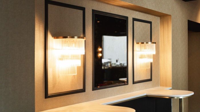 Detail foyer Theaterhotel De Oranjerie – twee glazen wandlampen met warme gloed naast een zwarte spiegel in panelenwand boven een afgeronde bar.