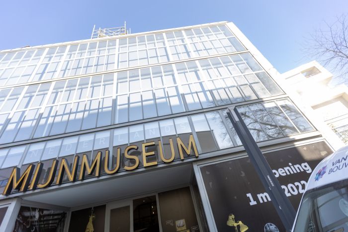 Glazen gevel van het Nederlands Mijnmuseum in Heerlen met grote gouden letters ‘MIJNMUSEUM’, steigerwerk bovenaan en raamstickers met ‘Opening 1 mei 2022’; rechts de neus van een bedrijfsbus.
