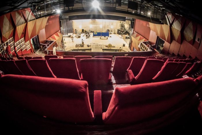 Uitzicht vanaf balkon: rode stoelen, open podium in opbouw met materialen en technici op de vloer